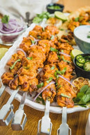Grilled chicken skewers made with the Napoleon Rotisserie Shish-Kebab Skewer Set (64008) by Napoleon Grills are garnished with herbs and red onion, served on a platter with lime wedges, sliced jalapeños, and a bowl of sauce in the background.