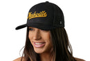 A woman smiles and looks to the side against a white background, wearing the Haka Hat Nashville Gold Script Trucker Hat— a black Tennessee snapback by Haka Hat.