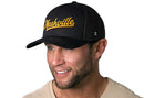 A smiling man wears the Haka Hat Nashville Gold Script Trucker Hat, a black Tennessee snapback by Haka Hat, paired with a light beige shirt against a white background.