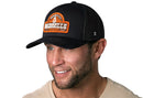 A smiling man wears the Haka Hat Nashville Music City Trucker Hat, a black cityscape snapback by Haka Hat with an orange and white "Nashville, Tennessee" patch, paired with a beige t-shirt against a white background.