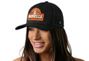 A woman with long brown hair smiles while wearing the Haka Hat Nashville Music City Trucker Hat—Black Cityscape Snapback. She stands against a plain white background.