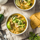 Two bowls of chicken chili with white beans, avocado, lime, cilantro, and a sprinkle of SALTOPIA Infused Sea Salts' CHEDDAR CHEESE salt, served with tortilla chips, jalapeño, fresh cilantro sprigs, and two spoons on a beige cloth.