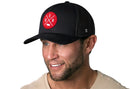 A smiling man wears the Haka Hat Nebraska Trucker Hat—black mesh snapback with a red NEB patch—paired with a beige shirt, against a white background.