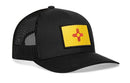 The Haka Hat New Mexico Flag Trucker Hat is a black mesh snapback featuring a yellow patch with a red Zia sun symbol on the front, representing the state flag.