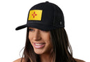 A woman with long brown hair smiles while wearing the Haka Hat New Mexico Flag Trucker Hat | Black NM Snapback, a black snapback by Haka Hat featuring a yellow patch with a red Zia sun symbol. The background is plain white.