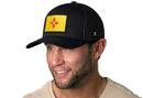 A smiling man in a tan shirt with light facial hair wears the Haka Hat New Mexico Flag Trucker Hat, a black NM snapback featuring a yellow patch with the red Zia sun symbol.