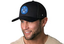 A smiling man with stubble wears the Haka Hat New York City Trucker Hat—a black NYC X Snapback with a blue and red logo. He pairs it with a light shirt and looks off to the side against a white background.