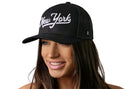 A woman with long brown hair smiles while wearing a Haka Hat New York City White Script Trucker Hat, a black snapback by Haka Hat featuring “New York” in white script embroidery. The background is plain white.