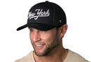 A smiling man with a trimmed beard wears a Haka Hat New York City White Script Trucker Hat in black and a light beige shirt, against a plain white background.