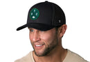 A smiling man wears a Haka Hat New York Trucker Hat—black, with a green circular NYC embroidered patch and black mesh back—paired with a light beige shirt. The background is plain white.