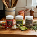 A Saltopia Flavor Trio Set – Hot Honey, Lime Garlic & Za’atar Infused Sea Salts arranged on a wooden counter with garlic bulbs, lime wedges, fresh herbs, and a honey dipper.