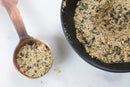 A black bowl and a copper measuring spoon, both filled with the light brown, coarse blend of World Spice's Americana Tasting Flight, showcase the aromatic spice mixes inspired by regional USA flavors as they rest on a white surface.