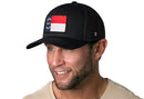 A smiling man in a beige shirt wears the Haka Hat North Carolina Flag Trucker Hat | Black NC Snapback, featuring a rectangular NC state flag patch on the front. The background is white.