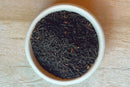 A white bowl filled with World Spice Northwest Tea Time loose black tea leaves sits on a light wooden surface.