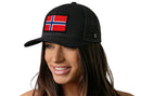 A woman with long brown hair smiles in a white background, wearing the Haka Hat Norwegian Flag Trucker Hat—a black snapback by Haka Hat featuring a Norway flag patch on the front.