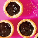 Three mini chocolate tarts with flaky crusts and glossy filling are topped with SALTOPIA Infused Sea Salts’ Saltopia Chocolate Sugar: HEYO COCOA 16oz, arranged on a bright pink background with scattered sea salt flakes.