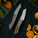 The Messermeister Ojai Damasteel Folder - 3.75" sits on a dark surface with oranges and green leaves, its eye-catching Damascus blade making it an excellent choice for everyday carry.