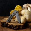 A Messermeister Ojai Folder - 3.75" with a grey handle sits open on a rustic wooden surface among yellow and white mushrooms. This everyday carry knife, crafted from N690 super steel, stands out against the dark background.