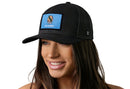 A smiling woman wears the Haka Hat Oklahoma Flag Trucker Hat— a black snapback by Haka Hat with a blue patch showing the Oklahoma state flag and emblem. The background is plain white.