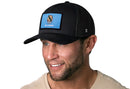A man smiles at the camera wearing a Haka Hat Oklahoma Flag Trucker Hat, featuring a black snapback and an Oklahoma flag patch on the front.