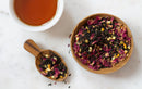 A bowl and a wooden spoon filled with World Spice Onyx Rose—a loose leaf tea blend from World Spice with Keemun tea, rose petals, and dried orange—sit next to a cup of brewed floral tea on a white marble surface.