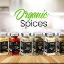 Twenty-four glass jars with sleek black labels from The Spice Lab’s Ultimate Organic Spice Set are neatly arranged on a wooden countertop, forming an elegant display. In the background, a bright kitchen adds warmth to the scene. Bold green and black text above the jars proclaims: “USDA Certified Organic Spices.”.