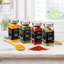 Four glass spice jars labeled Ground Turmeric, Parsley Flakes, Spanish Sweet Paprika, and Bay Leaf from The Spice Lab Organic Starter Spice Set - 18 Jars are arranged on a white table with a wooden spoon, spice powder, and bay leaf. This USDA Certified Organic set by The Spice Lab is displayed in a bright, airy kitchen background.