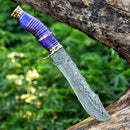 The Texan Knives Vintage Original Damascus Steel Bowie Knife – 13" with brass guard and camel bone handle is displayed on a tree branch, with blurred green foliage in the background.
