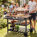 A man grills meat on the DirectDeals Outdoor Dining Cart with BBQ Grill and Folding Side Panel, while friends enjoy food and laughter at an outdoor table in a sunny garden.