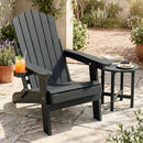 A black DirectDeals Folding Adirondack Chair with Adjustable Backrest & Cup Holder sits on a stone patio beside a dessert table, holding an orange drink with lime. Greenery and potted plants surround the sunny outdoor space.