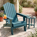 A Folding Adirondack Chair with Adjustable Backrest & Cup Holder by DirectDeals sits on a stone patio beside a table with a drink, book, and dessert. Sunlight and greenery add to the inviting outdoor scene.