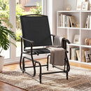 The DirectDeals Outdoor Glider Chair with Metal Frame and Armrests sits on a rug in a bright room, draped with a gray blanket. Sunlight streams in near bookshelves, decor, and a potted plant.