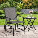 A DirectDeals Outdoor Glider Chair with Metal Frame and Armrests faces a round table holding pink flowers, water, and a newspaper on a rug outside, surrounded by grass and trees.