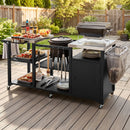 A DirectDeals Outdoor Grill Cart with Stainless Steel Tabletop & Cabinet sits on a deck holding a grill, drinks, plates, utensils, food, and a trash bag, against a backdrop of a fenced backyard with greenery.