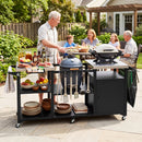 An older man grills food at a backyard DirectDeals Outdoor Grill Cart with Stainless Steel Tabletop & Cabinet, while adults and a child share a sunny meal. Plates, utensils, and fresh food are neatly arranged on the stainless cart.
