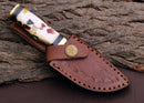The Texan Knives Custom Handmade Damascus Steel Outdoor Knife features a 3" blade and a multicolored handle, secured in a brown leather sheath with decorative stitching and a brass emblem button, resting on textured bark.
