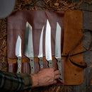 A hand reaches for one of five knives with textured handles, arranged on a Messermeister Overland Leather 5 Pocket Knife Roll With Pin Closure in Cognac Brown by Messermeister, displayed on wood among scattered dried leaves.
