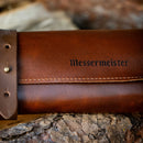 A close-up of the Messermeister Overland Leather 5 Pocket Knife Roll With Pin Closure in Cognac Brown rests on rough tree bark, featuring embossed Messermeister branding.