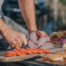 Outdoors, someone slices a red onion on a wooden cutting board with the Messermeister Overland Chef Camp Knife - 8", surrounded by sliced tomatoes and bread rolls. Sunlight highlights the fresh ingredients and their hands as they prepare food.