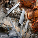 The Messermeister Overland Chef Scalloped Slicer - 8" is displayed among other knives on a beach-inspired setting with rocks, seashells, crabs, and a wooden board, evoking a coastal culinary adventure.