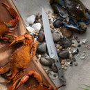 A Messermeister Overland Chef 6-Piece Leather Knife Roll Set rests on rocks and shells beside seasoned cooked crabs on a wooden board and a live blue crab on sand—creating the perfect scene for lovers of outdoor cooking knives.