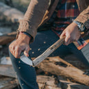 Wearing a brown jacket and plaid shirt, a person uses the Messermeister Overland Chef Utility Knife - 4.5" to carve wood, sending chips flying from the stick.