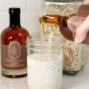 A hand pours pure maple syrup into a mason jar of oats and milk. Nearby, a large jar of oats and a bottle of Under The Bark Syrup Co Coffee Maple Syrup sit on the counter.