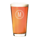 A Pint Glasses - Set of 4: The Medalist personalized pint glass filled with amber beer topped with foam, featuring a circular emblem with the letter M etched on the front.