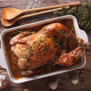A golden-brown roast chicken garnished with fresh herbs and SALTOPIA Infused Sea Salts’ Herbs De Provence Seasoning Salt in a white baking dish, surrounded by garlic cloves, thyme, wooden spoons, and sprinkled salt on a rustic wooden table.