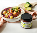 A rice bowl with shrimp, pineapple, jalapeños, salsa, and red cabbage sits beside a jar of Terrapin Ridge Farms Pineapple Chile Lime Salsa by Terrapin Ridge Farms. Lime wedges and jalapeños are on a cutting board nearby.