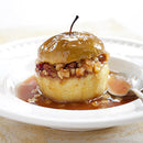 A whole baked apple filled with chopped nuts is topped with a glossy SALTOPIA Infused Sea Salts BEAN GOOD Vanilla Bean Sugar sauce, served on a white plate with caramel sauce and a spoon.