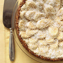 A pie topped with banana slices and coconut sits on a yellow cloth beside a metal server, its sweetness elevated by a dusting of SALTOPIA Infused Sea Salts’ Saltopia Vanilla Bean Sugar: BEAN GOOD 16oz.