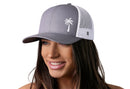 A woman smiles wearing the Haka Hat Palm Tree Trucker Hat, a gray and white Island Snapback with a palm tree logo by Haka Hat.