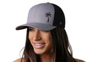 A woman with long brown hair smiles while wearing the Haka Hat Palm Tree Trucker Hat, a gray-black island snapback featuring a black logo on the front. The background is white.